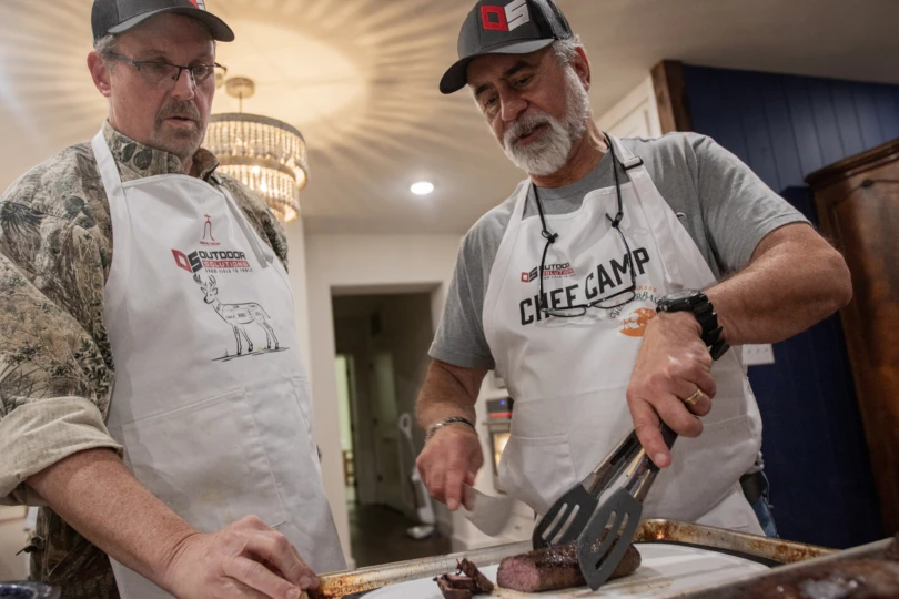 A chef and a student cutting a deer steak in a kitchen.