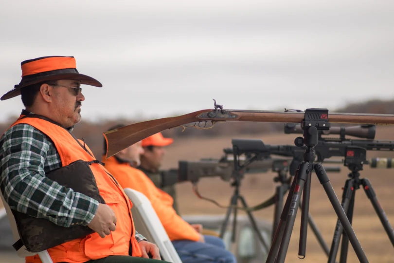 A hunter leaned back in a chair on a shooting range while his old flintlock muzzleloader rests on a shooting tripod.