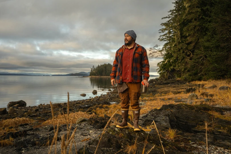 A man stands near the water holding a metal cup on an Alaskan shoreline