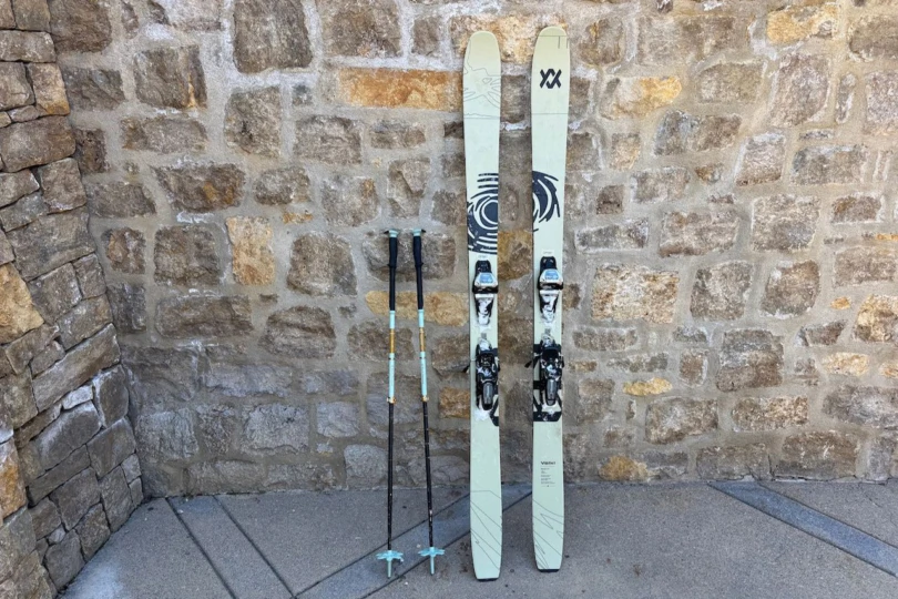 The Völkl Revolt 101 skis stand upright next to ski poles against a stone wall
