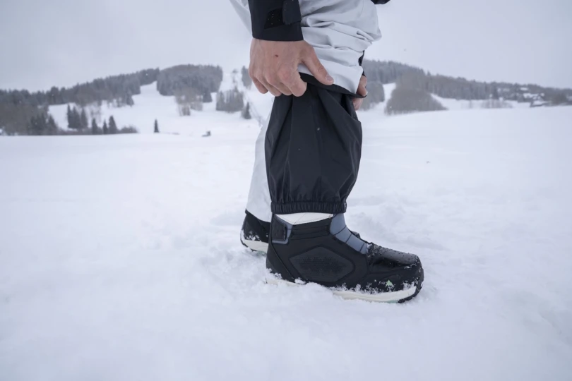 A person standing in deep snow adjusts their pant gaiters over winter boots