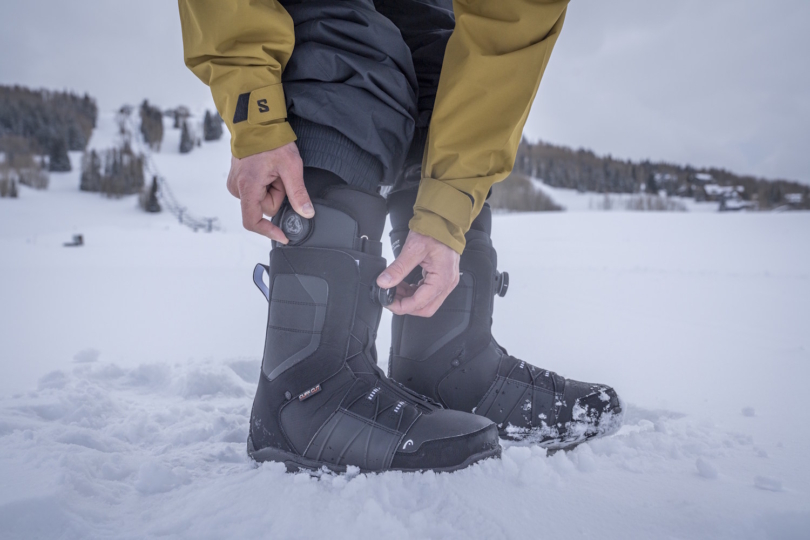 Snowboarder tightening BOA dials on black snowboard boots while standing in the snow