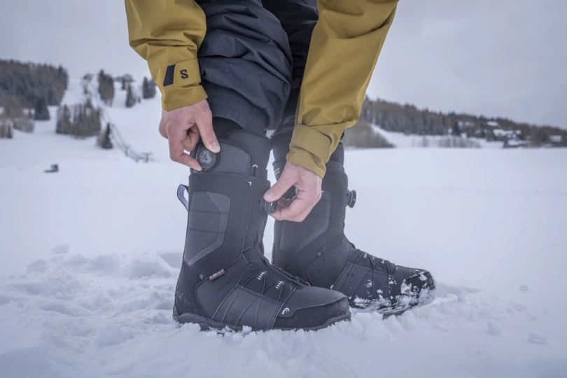 Snowboarder tightening BOA dials on black snowboard boots while standing in the snow