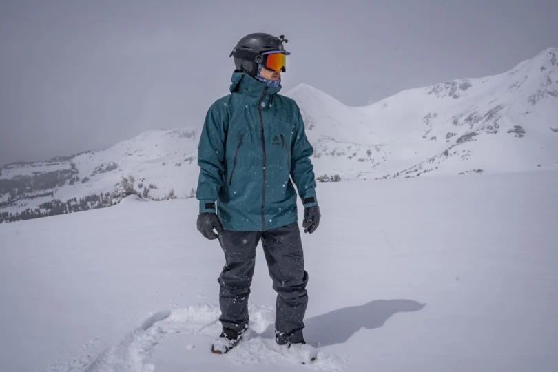 person in a teal snow jacket standing in deep snow with mountains in the background
