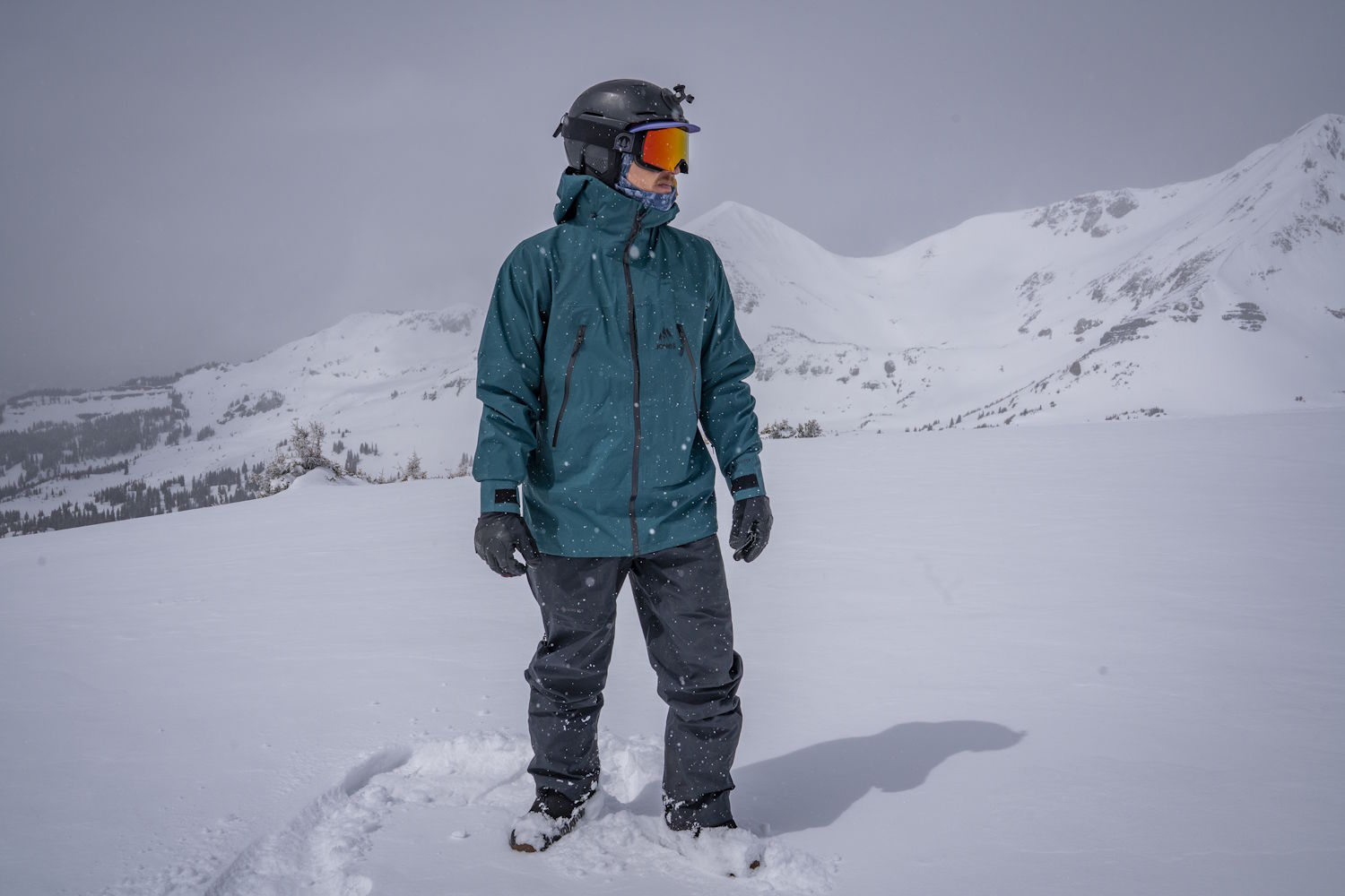 person in a teal snow jacket standing in deep snow with mountains in the background
