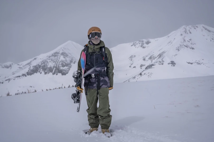 Snowboarder standing in deep snow with a board in hand, surrounded by snowy mountain peaks