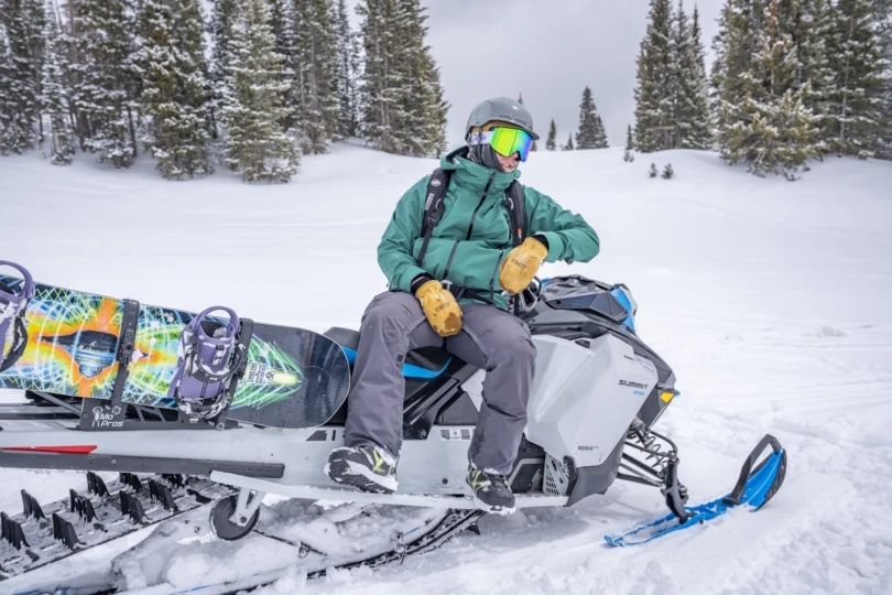 A person in winter gear sits on a snowmobile with a snowboard attached,