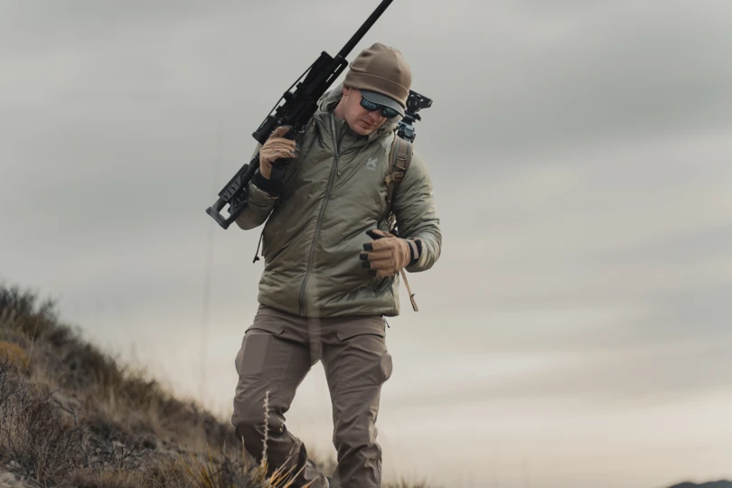 A hunter carrying a rifle moving on a steep slope while wearing the Kifaru Pointe Synthetic Puffy