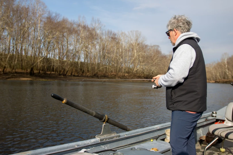 An angler fishing off the side of a small boat in a river.
