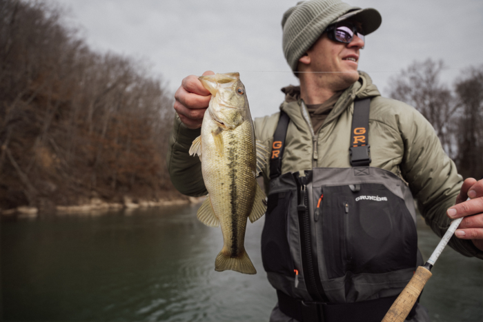 An angler wearing the Grundens Vector Zip wader holding a bass and a fly rod in front of a river.