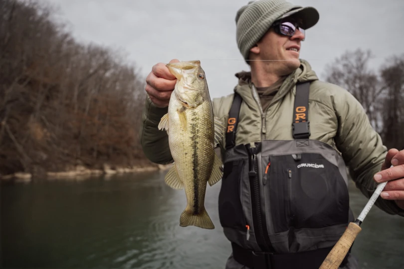 An angler wearing the Grundens Vector Zip wader holding a bass and a fly rod in front of a river.