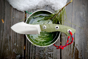 The Kizer Rook knife placed across a metal cup filled with water and pine needles