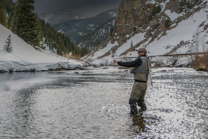 An angler stands midstream in a snowy mountain river wearing insulated layers while fly fishing
