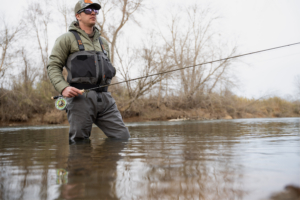 An angler wearing the Grundens Vector Zip waders standing in a river holding a fly rod.