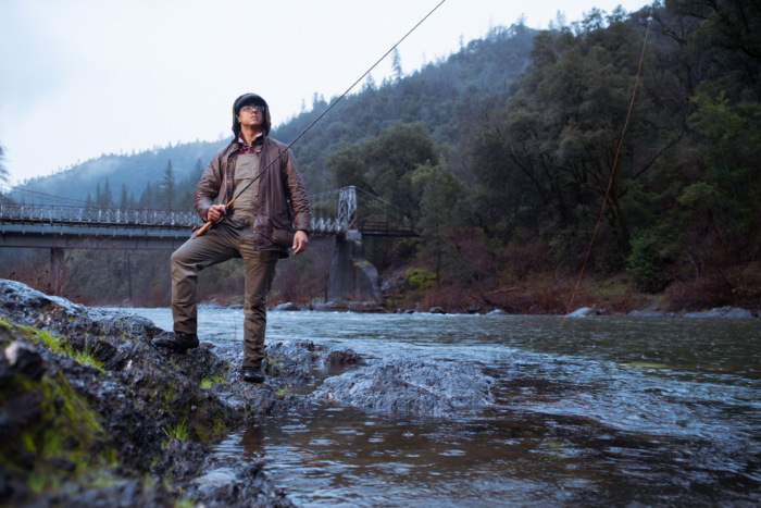 A person stands on wet rocks by a river while fishing, wearing the Barbour Beaufort Wax Jacket in rainy conditions