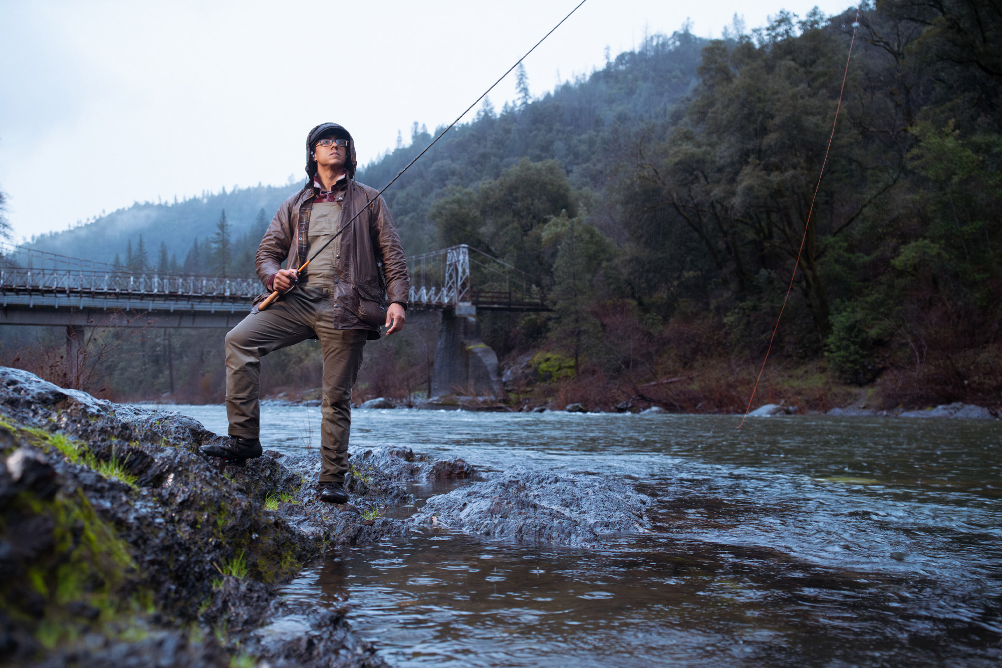 A person stands on wet rocks by a river while fishing, wearing the Barbour Beaufort Wax Jacket in rainy conditions