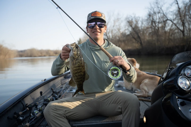 An angler holding a smallmouth bass and the Hardy Marksman fly rod on the bow of a motor boat.