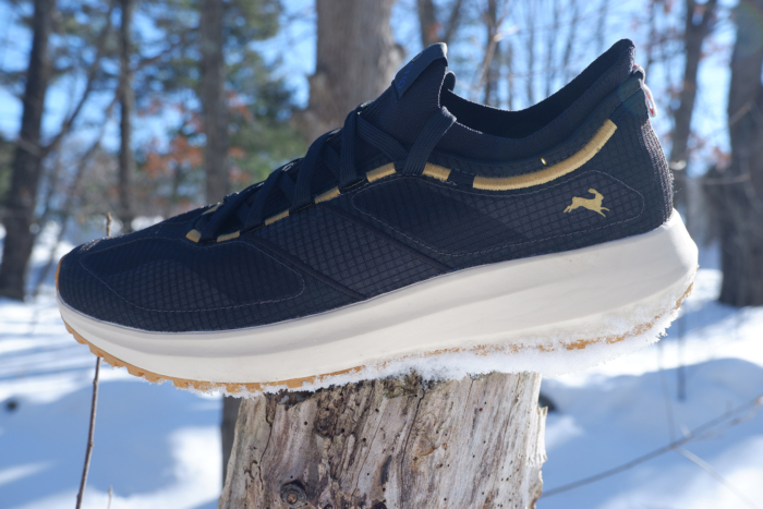 Side view of a Tracksmith Eliot NDO shoe balanced on a tree stump in a snowy forest