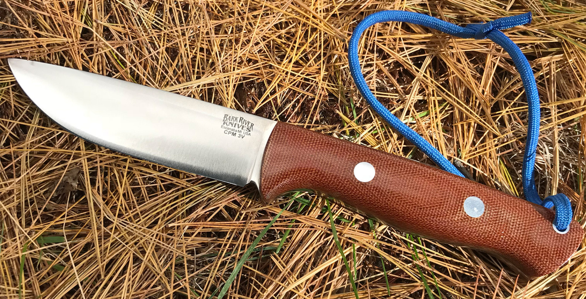 A Bark River fixed blade knife with a brown handle lies on dry pine needles with a blue cord attached