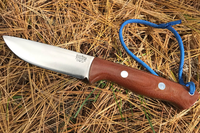A Bark River fixed blade knife with a brown handle lies on dry pine needles with a blue cord attached