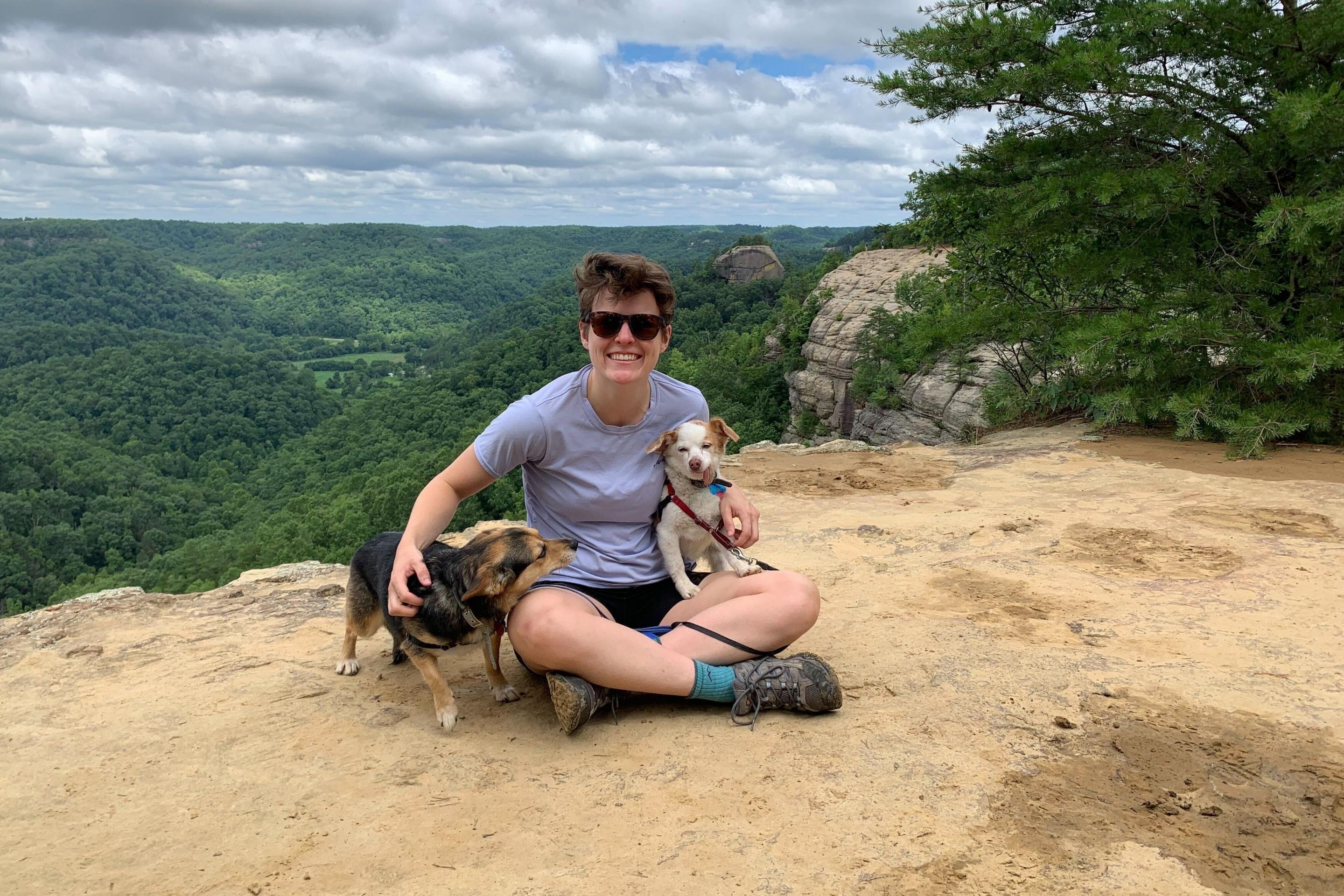 woman sits on cliffside with two dogs