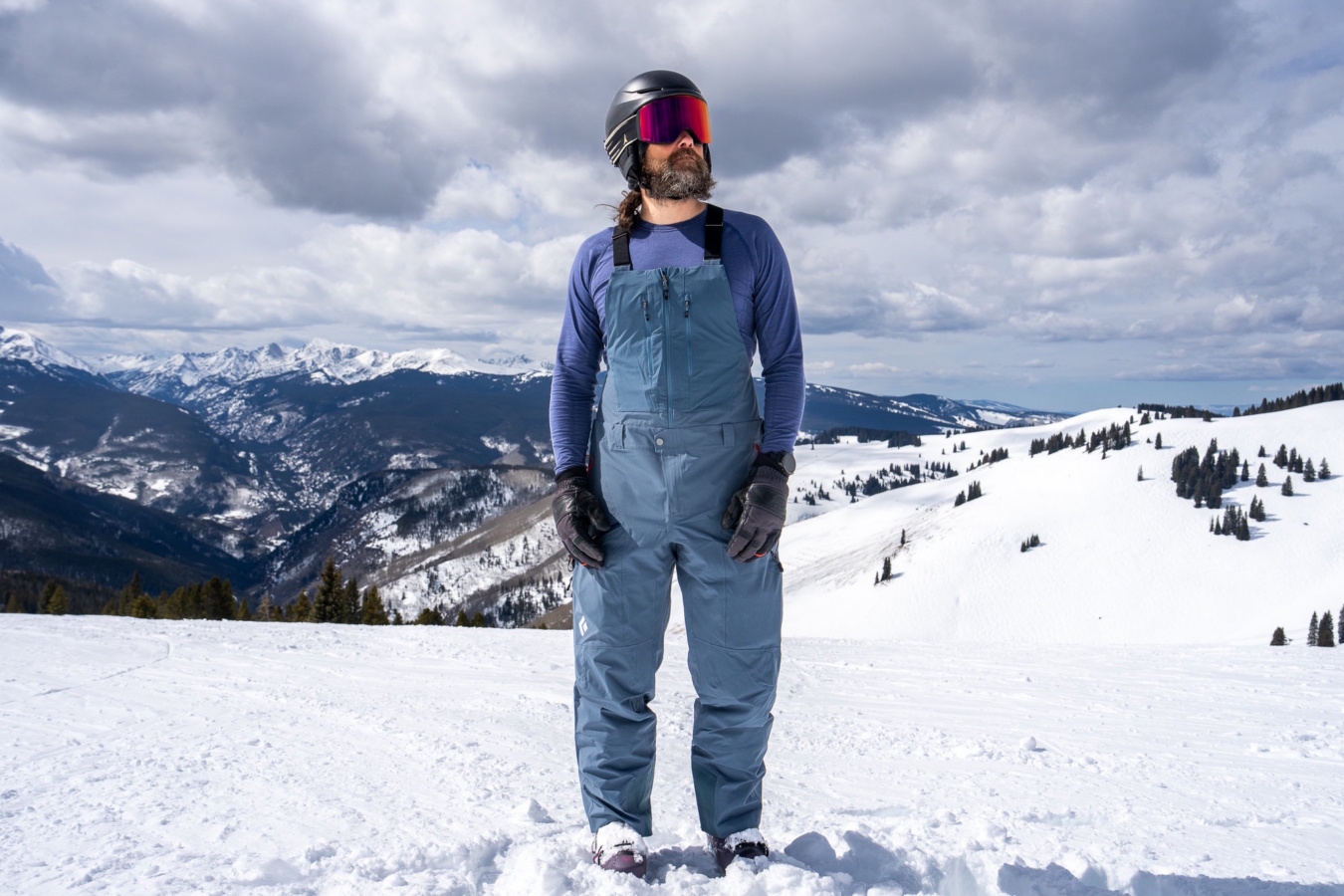 Skier wearing blue Black Diamond Recon Stretch Bibs standing on a snowy mountain ridge