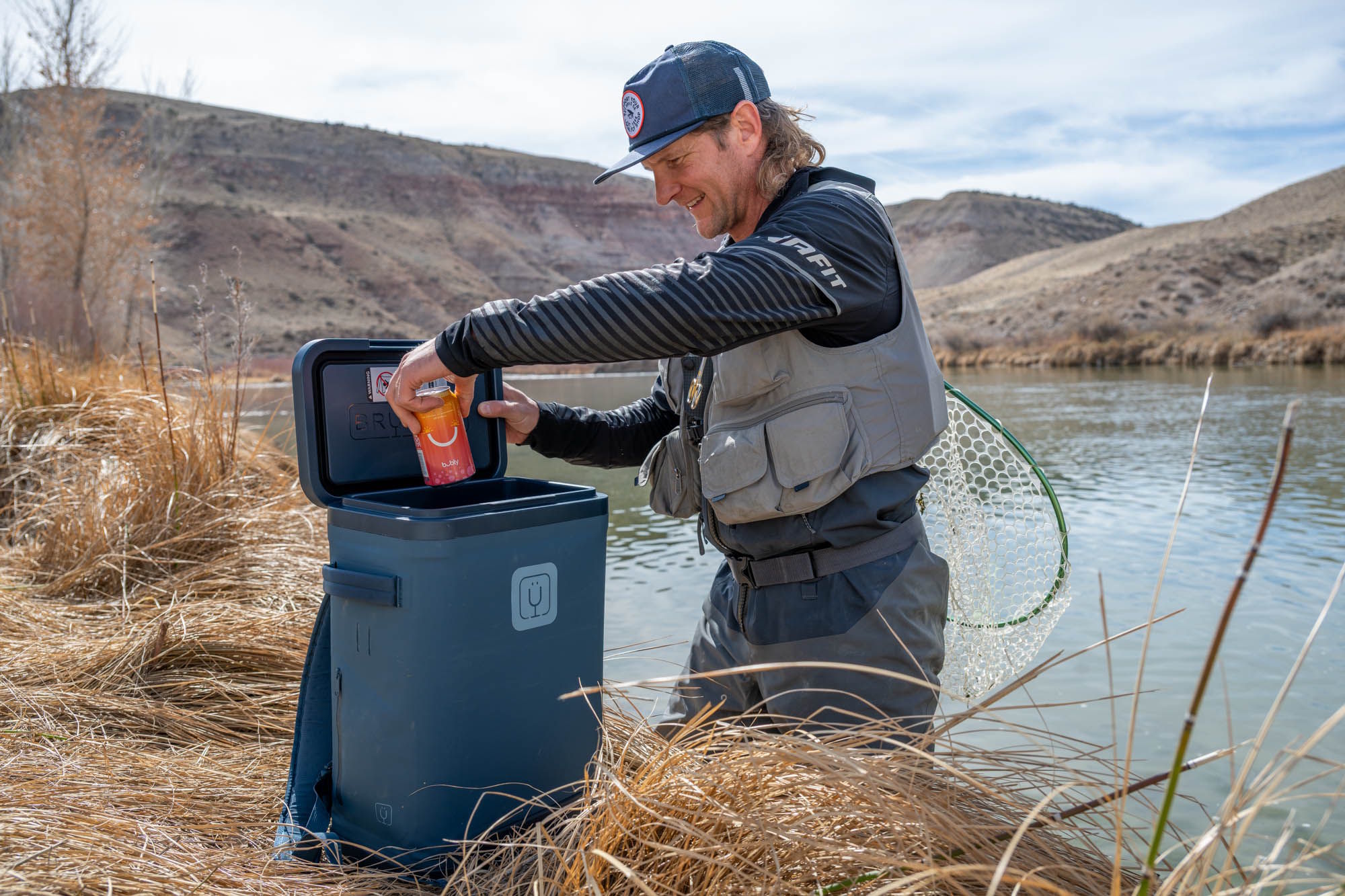 A man in fishing gear reaching into the open MagPack cooler by a river.