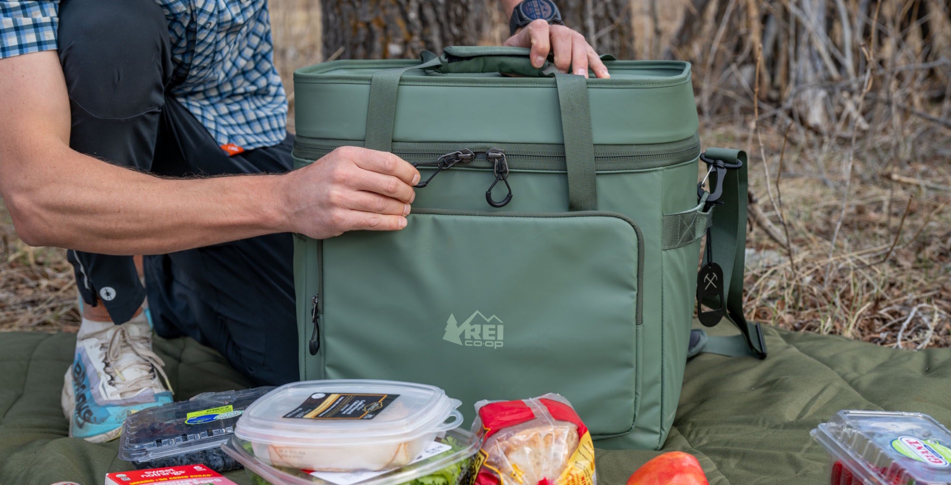 Person opening the REI Co-op Trailgate Cooler outdoors with fresh food and drinks arranged on a mat