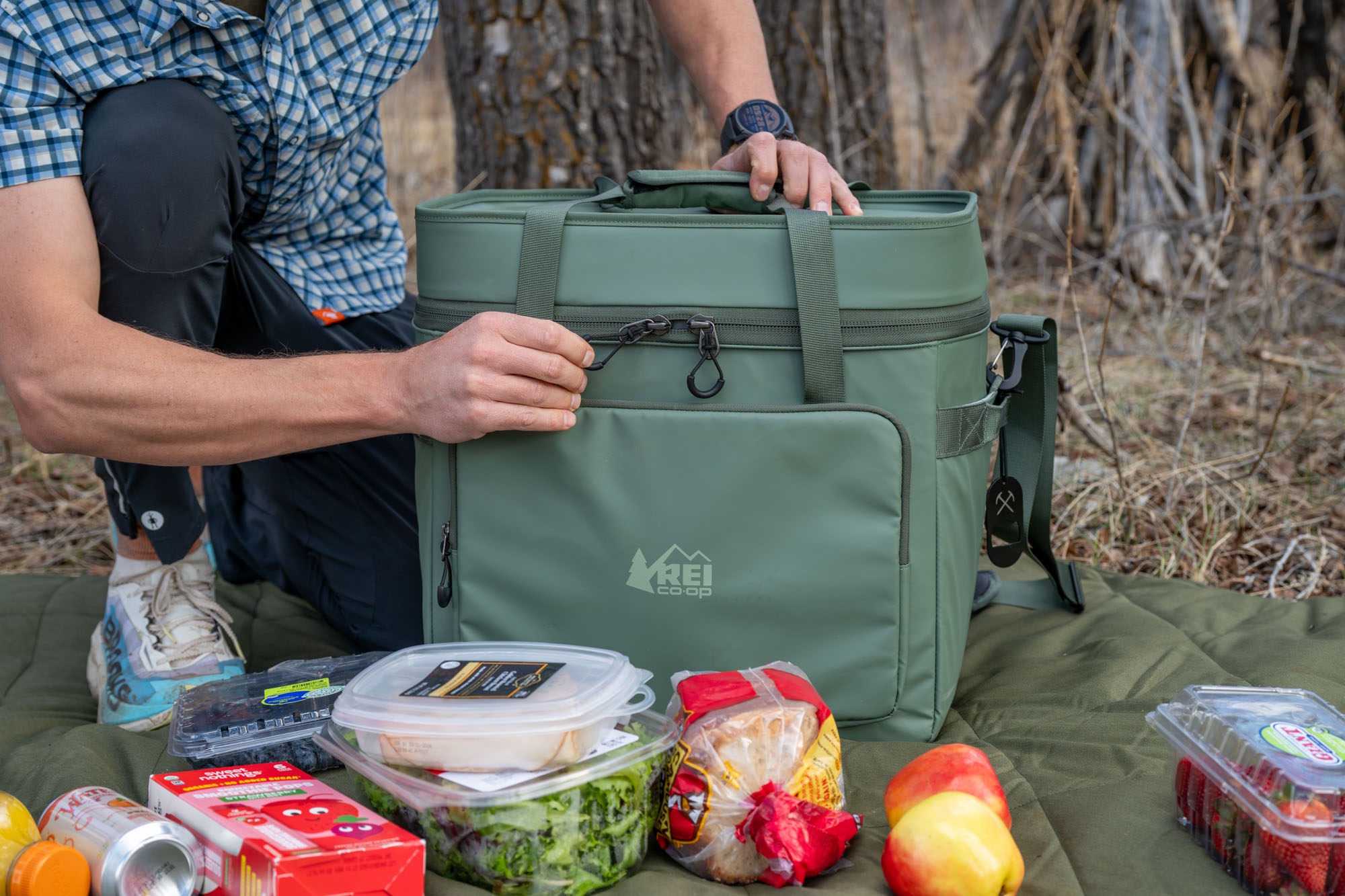 Person opening the REI Co-op Trailgate Cooler outdoors with fresh food and drinks arranged on a mat