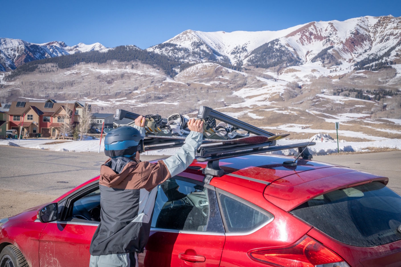 Person in a helmet loading skis onto a roof rack on a red car with snowy mountains