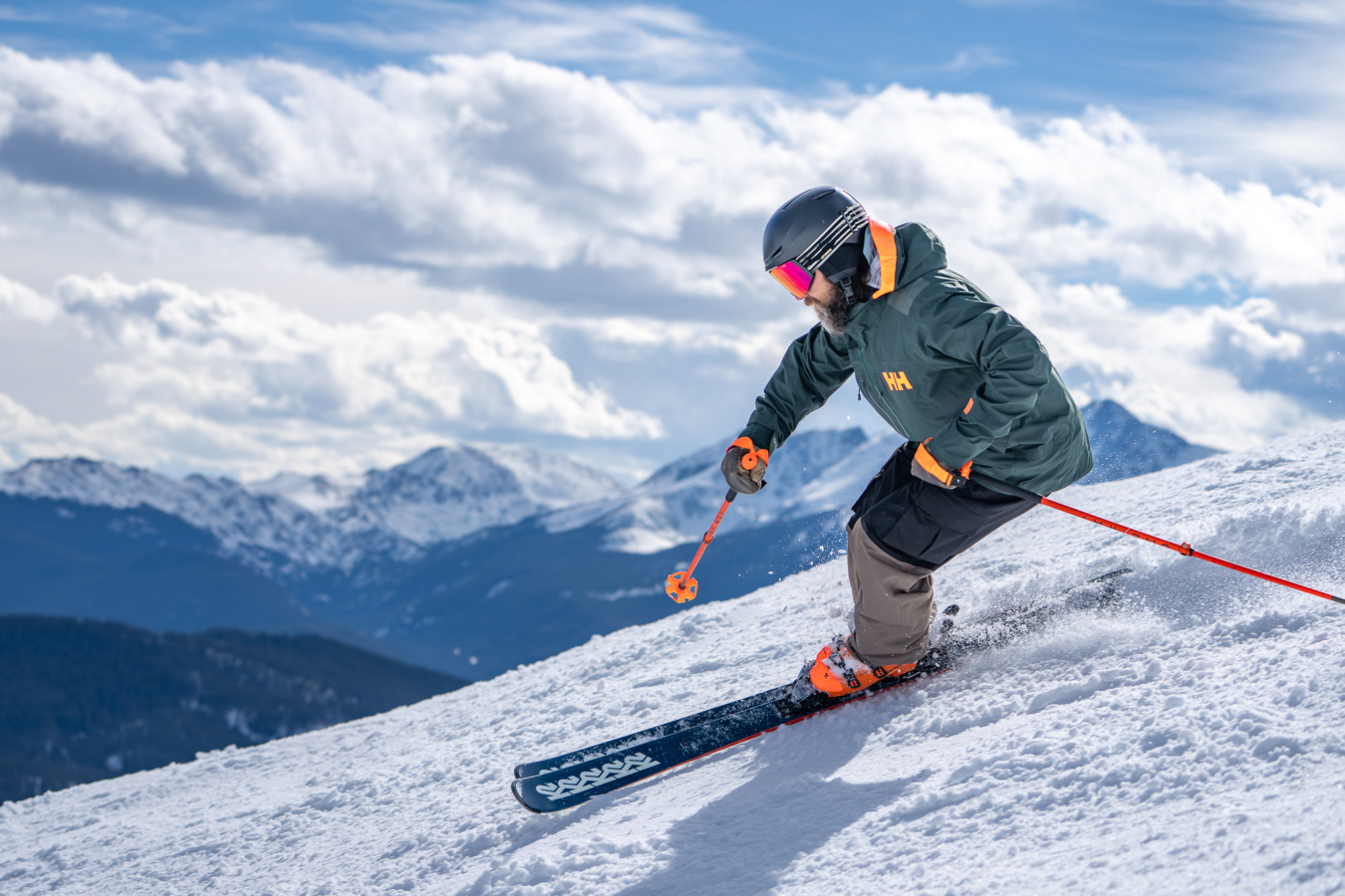 Skier wearing the Helly Hansen Ridge Infinity Jacket 2.0 skis across a snowy slope with mountains in the background