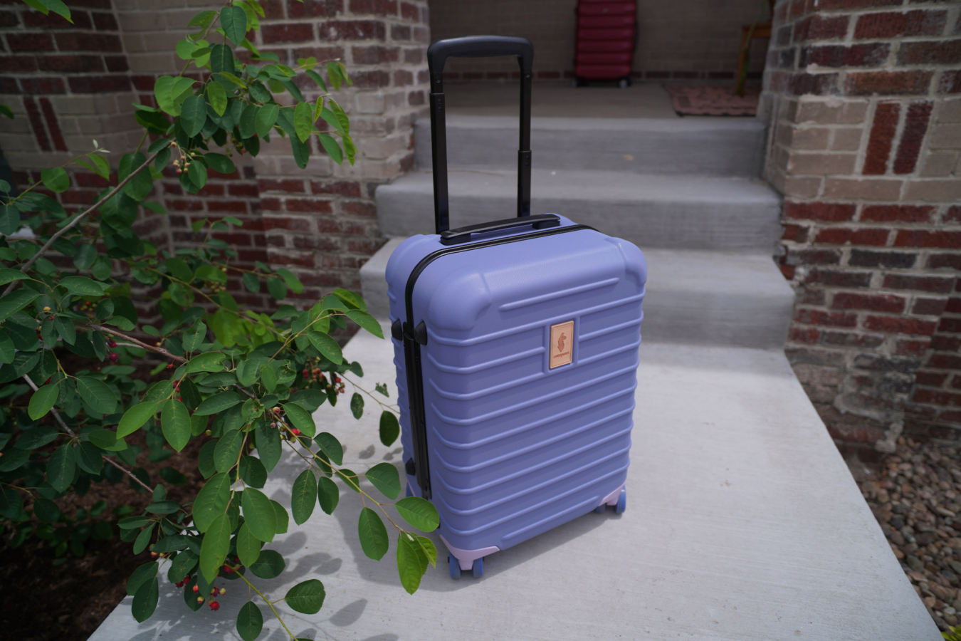 Purple Cotopaxi Coraza suitcase with the handle extended on a sidewalk near brick steps and greenery