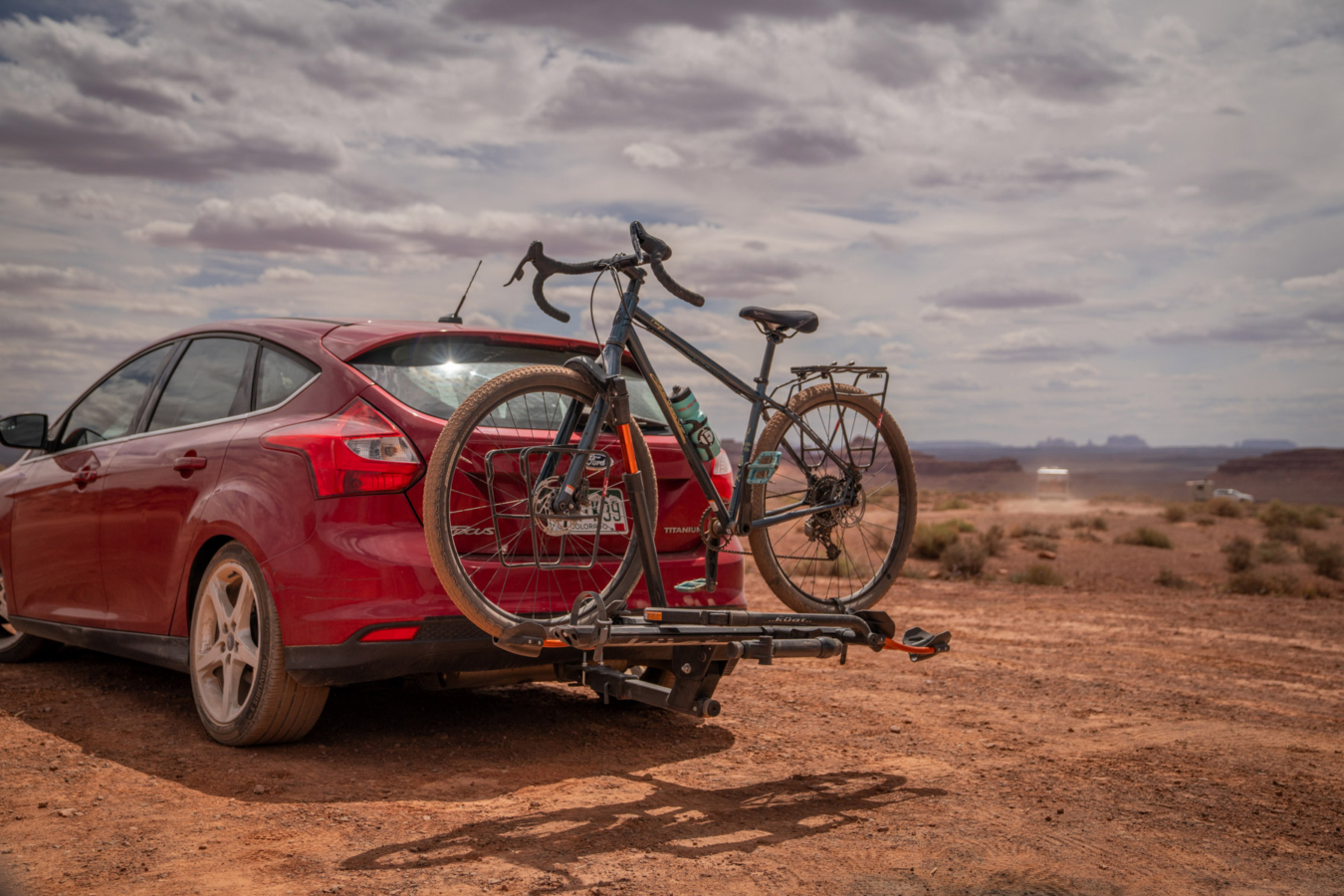 Bike mounted on a Kuat hitch rack behind a red car on a desert dirt road