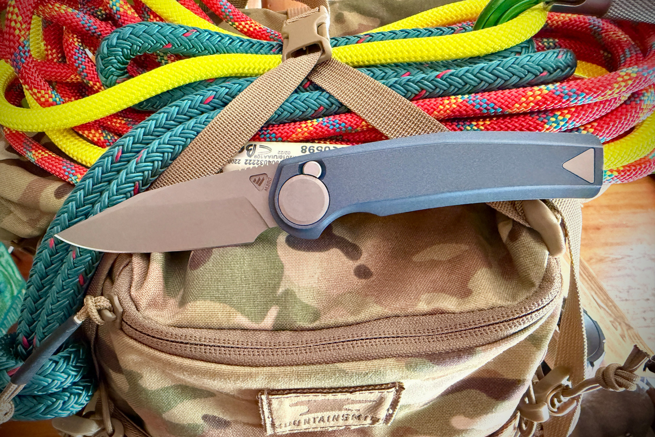 Open Madrone knife resting on a camouflage gear bag with colorful ropes behind it