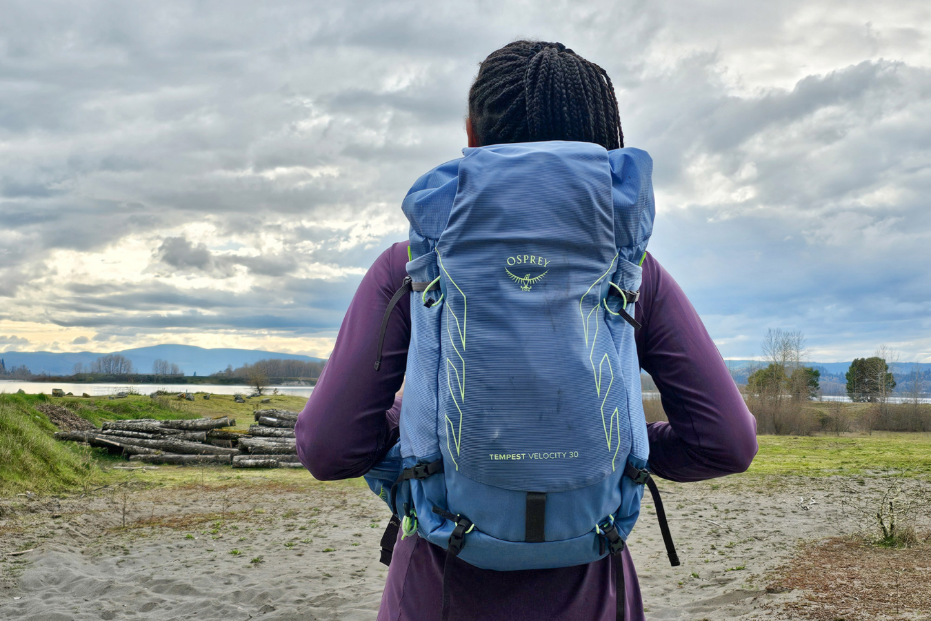 A hiker wears a blue Osprey pack near the water under a cloudy sky