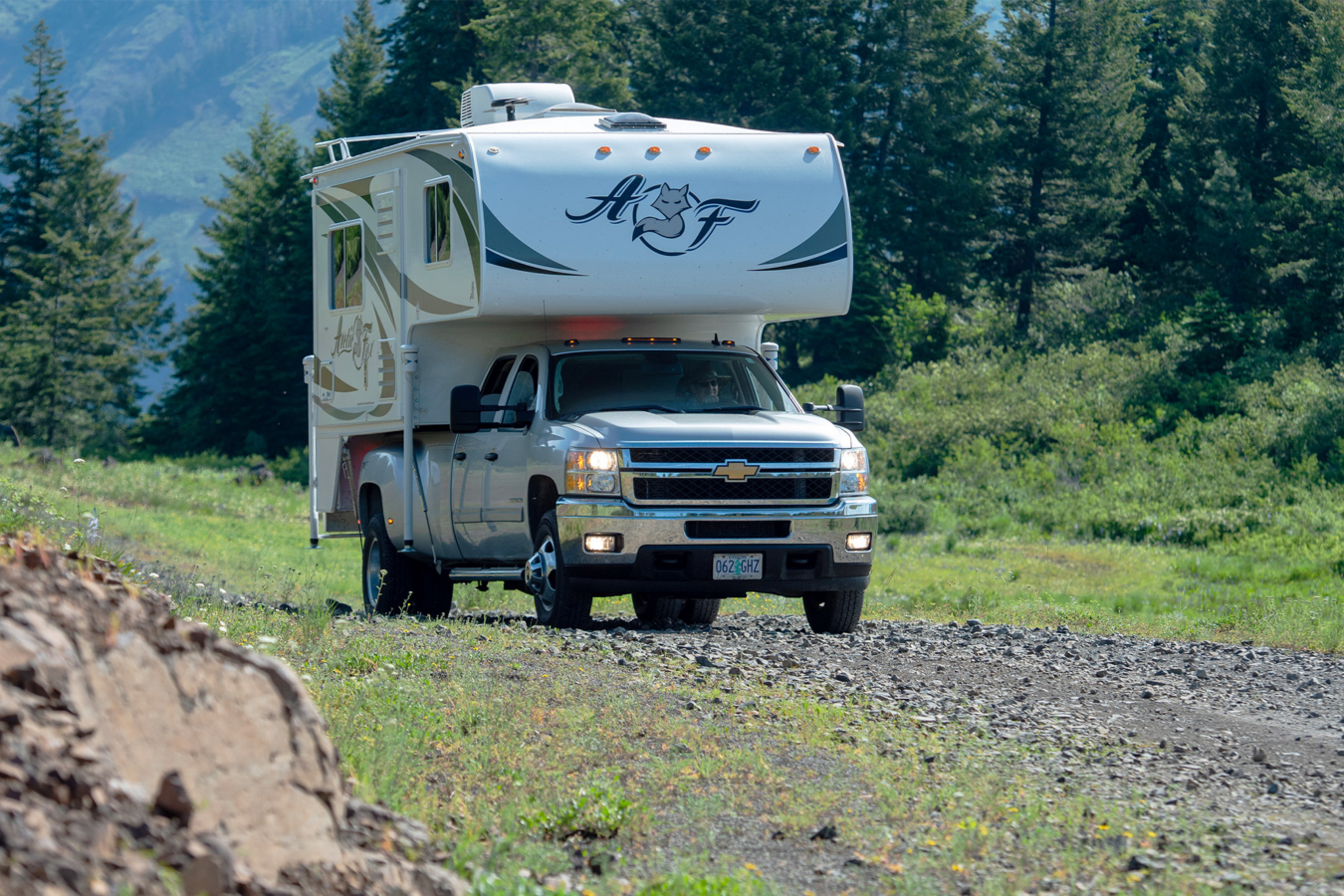 A pickup camper driving on a rocky dirt road through a forested mountain area