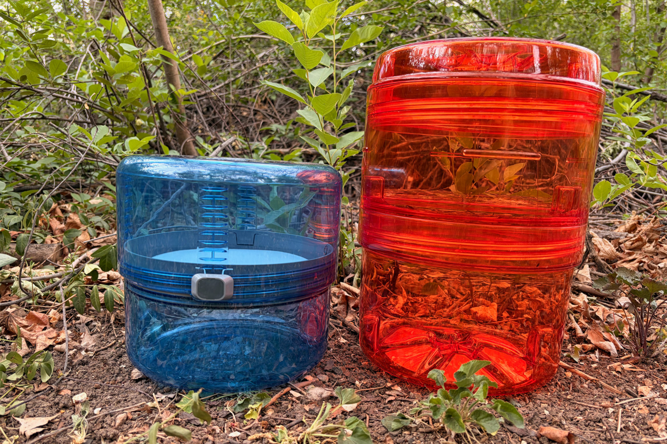 A blue BV ONE canister sits beside an orange REI Traverse canister on the ground in a wooded area