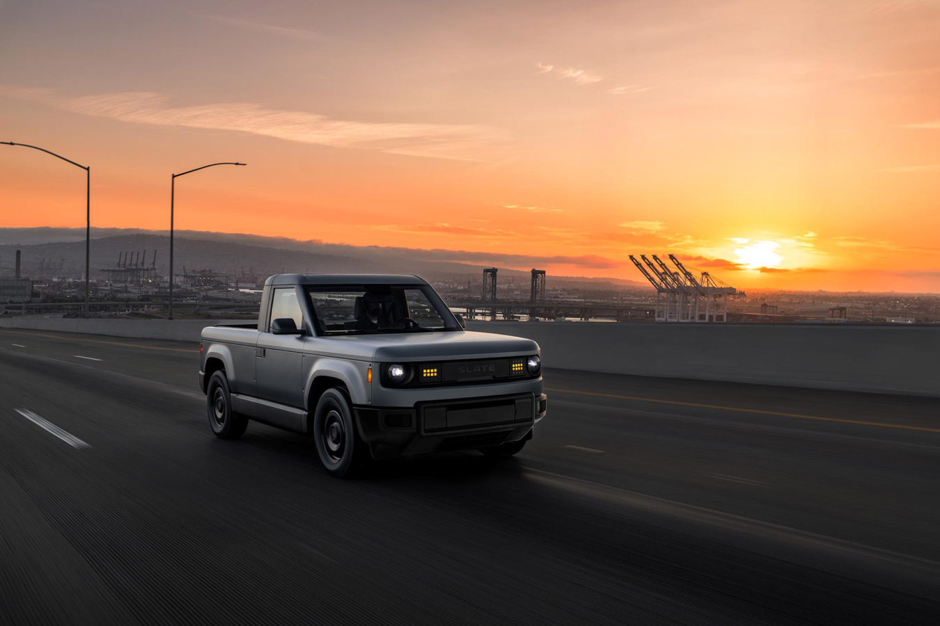 Slate pickup driving on a highway at sunset with port cranes in the background