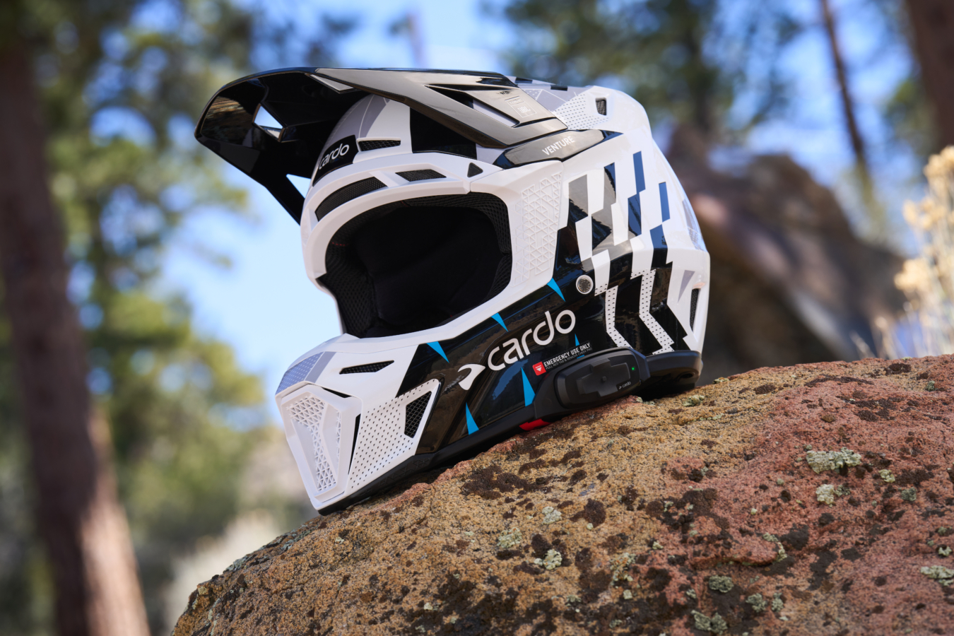 A white and black Cardo Venture dirt bike helmet sits on a rock,.