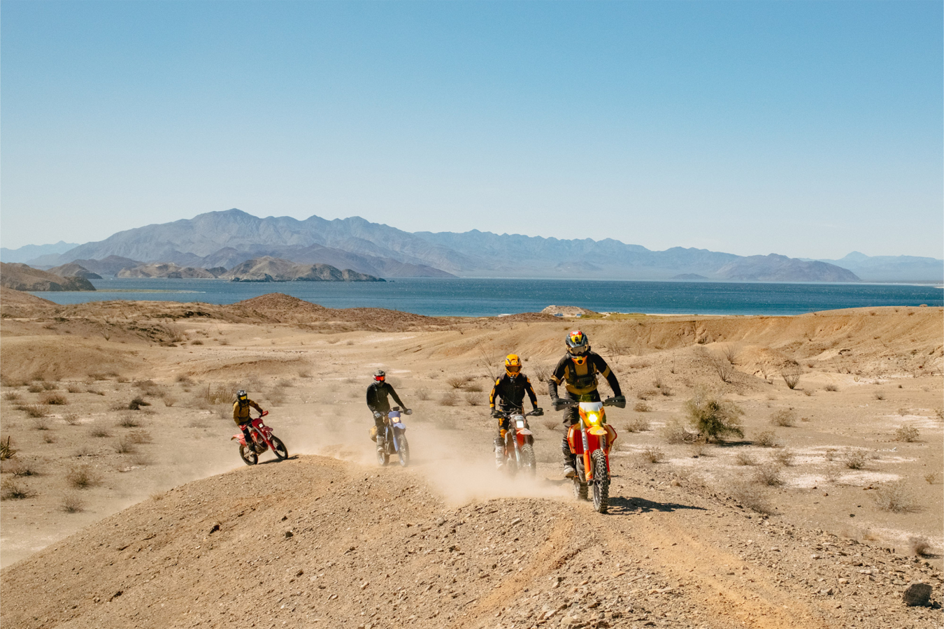 Four dirt bike riders crossing a desert trail near the Baja coast