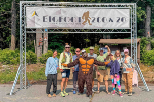 Runner and crew posing beneath the Bigfoot 200 Mile Endurance Run banner with a Bigfoot mascot