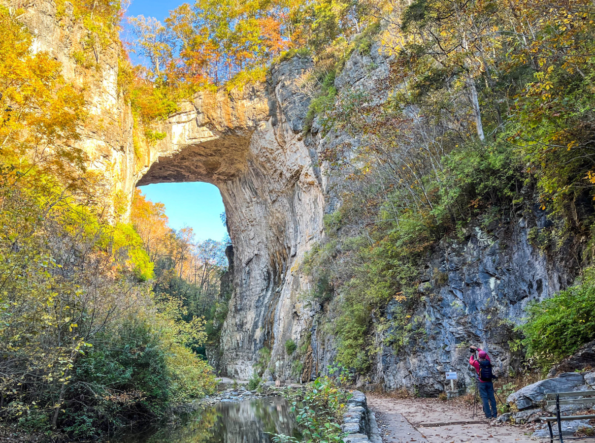 Hiker looks up at natural rock bridge surrounded by fall colors