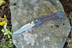 Buck Knives 110 Crossbar Elite folding knife with an open blade and wood handle resting on a mossy rock