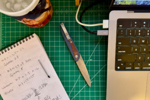 Open folding knife with a blue handle and silver blade resting on a green cutting mat beside a notebook and laptop
