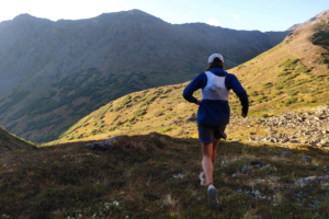 Runner wearing a white vest over a blue jacket while heading uphill on rocky terrain
