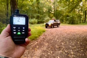 Hand holding a Midland radio handset displaying channel 2 with an off road SUV on a wooded trail in the background
