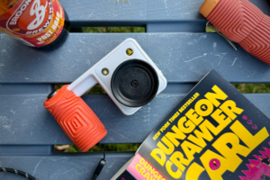 White NOCS Provisions Portal AFN7 camera with an orange grip resting on a blue picnic table beside a book and drink