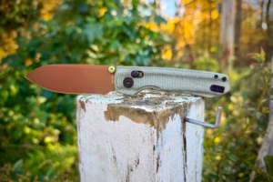 Open folding knife with a brown blade and green textured handle resting on a weathered wooden post