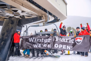 skiers exiting a ski lift with large banner reading "Winter Park. We're Open!"