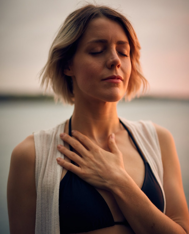 A woman in a black swim suit top and their eyes closed holding their hand across their heart